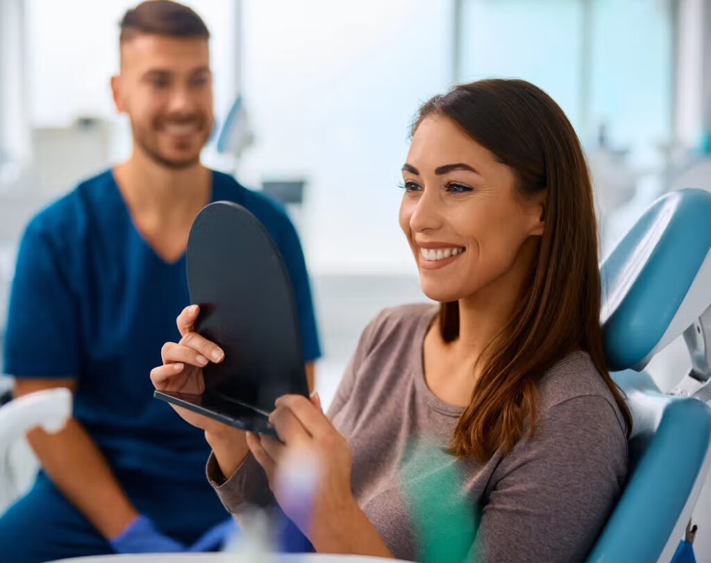 Emergency Dentist In New Port Richey: What To Do Right Now For A Toothache, Chip, Or Abscess Happy woman in a dental chair looking at her smile in a mirror, with a dental professional smiling nearby.