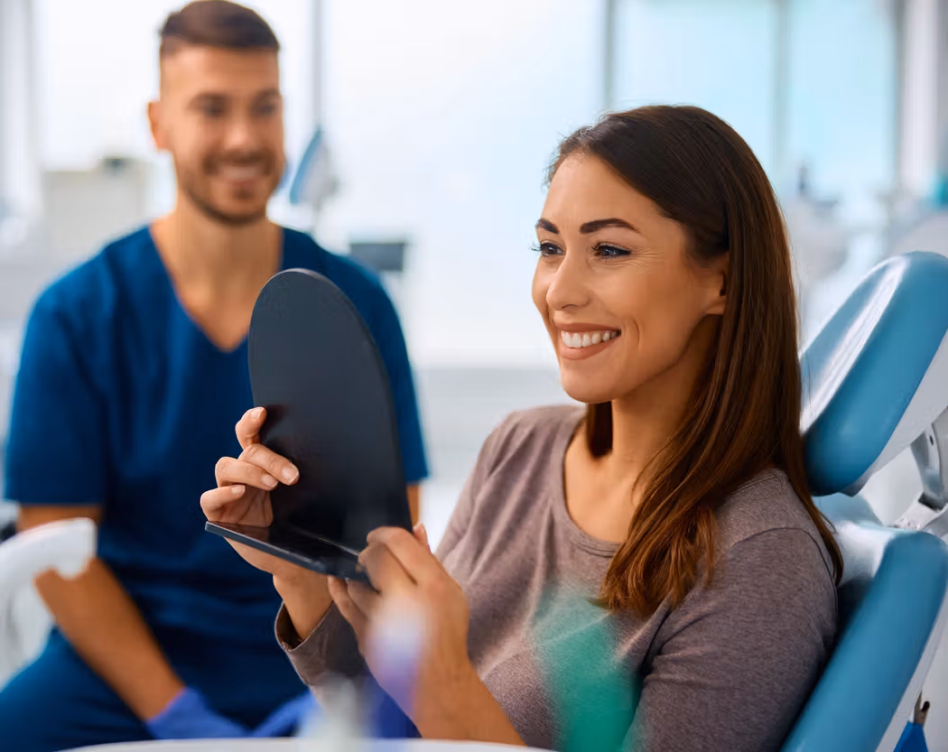 Happy woman in a dental chair looking at her smile in a mirror, with a dental professional smiling nearby.