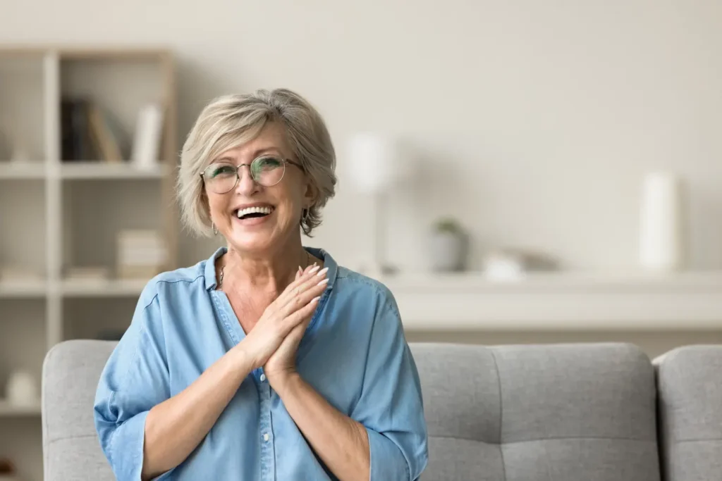 Implant Supported Dentures In Trinity: More Stability, Less Worry—Every Single Day A happy woman with glasses, hands clasped, sitting on a couch.