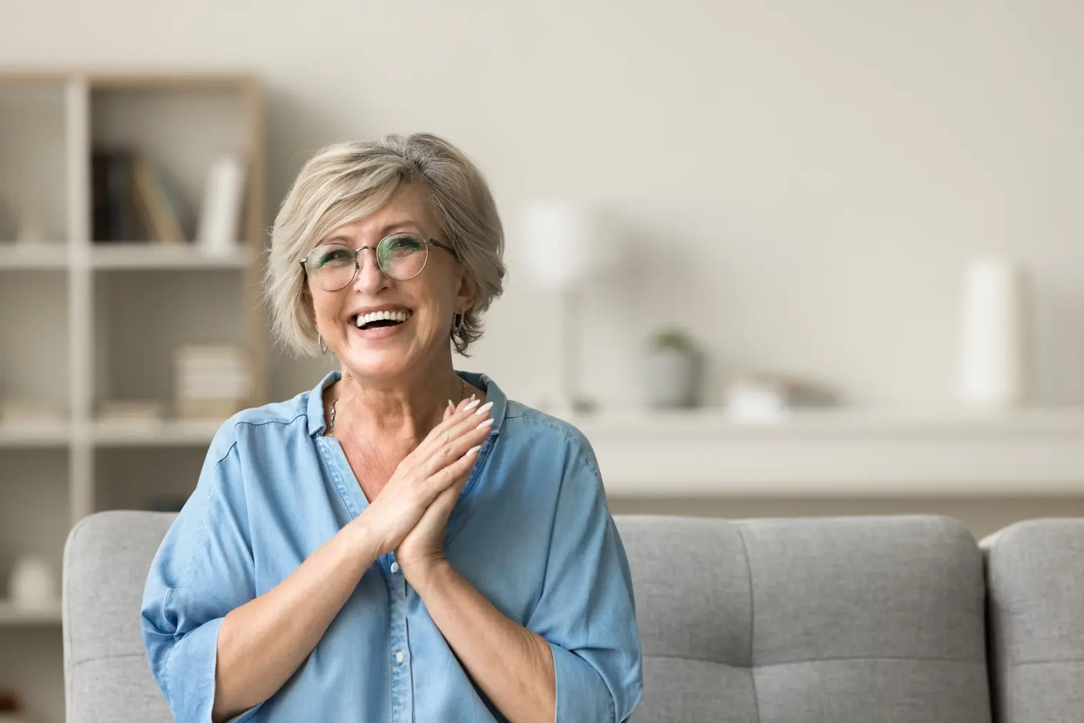 A happy woman with glasses, hands clasped, sitting on a couch.