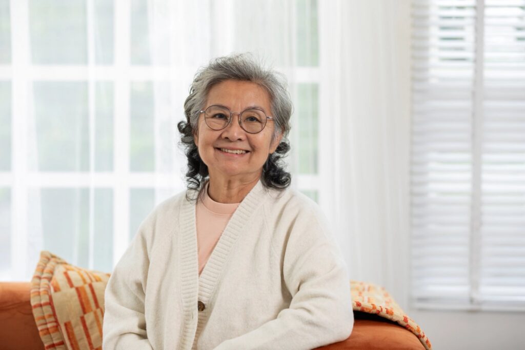Modern Dentures And Partials In Trinity: Comfort, Fit, And Options Smiling elderly Asian woman with glasses and gray hair sitting on a sofa.