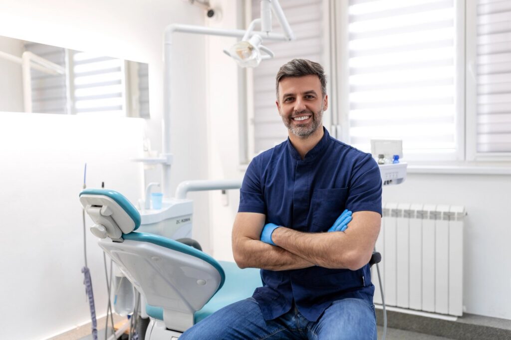 A smiling male dentist in blue scrubs and gloves sits in a modern dental office.