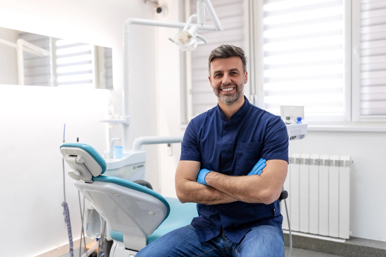 A smiling male dentist in blue scrubs and gloves sits in a modern dental office.