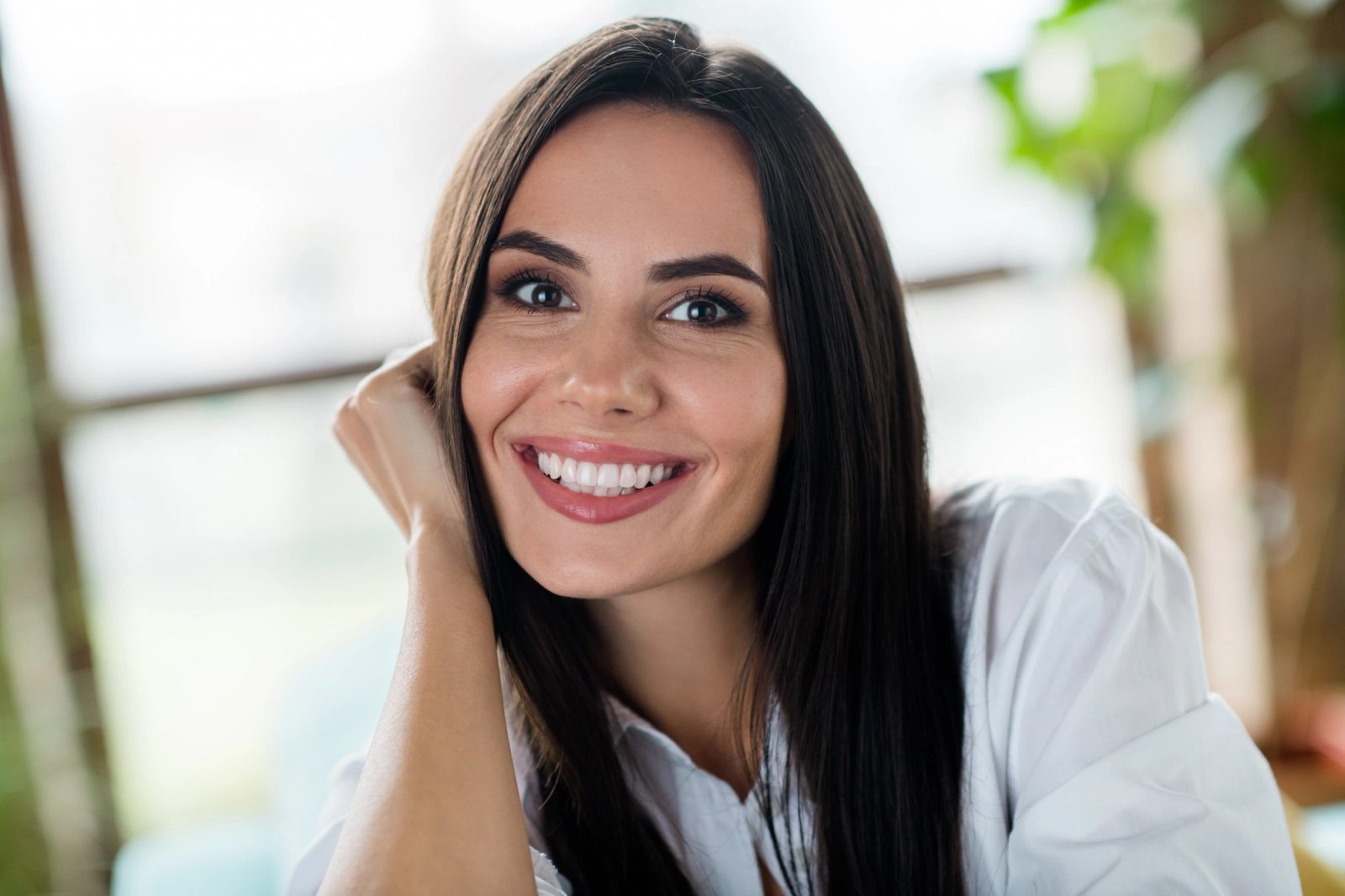 A smiling woman with long dark hair, wearing a white shirt, resting her hand by her face.