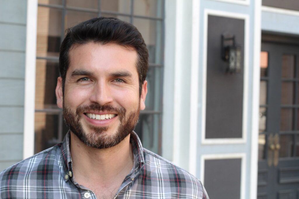 All-On-X Fixed Implants In Trinity: A Confident, Full-Arch Smile Without Slipping A smiling man with a beard and plaid shirt stands in front of a house.