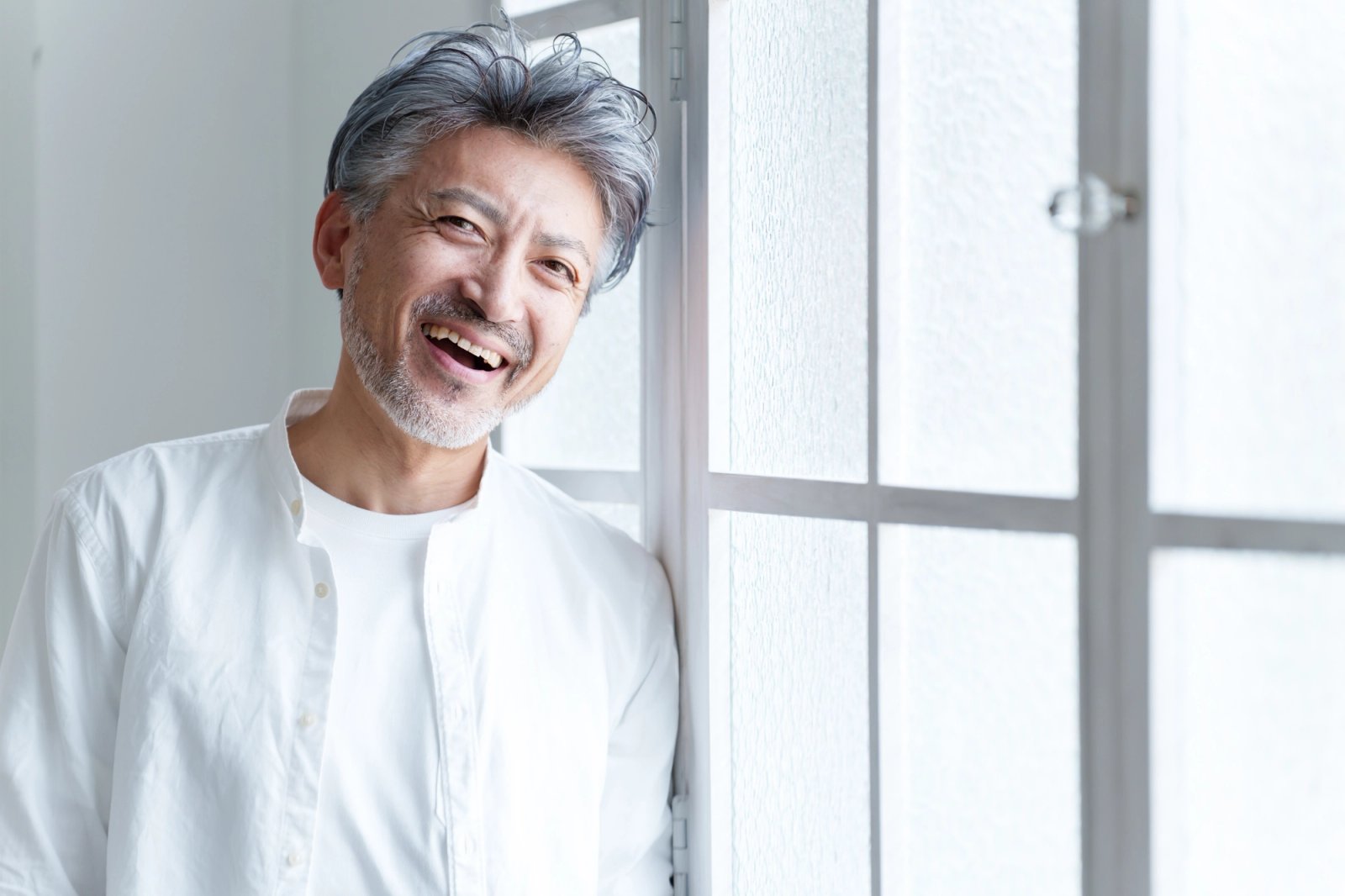 A man with gray hair and a beard laughing and leaning against a window.