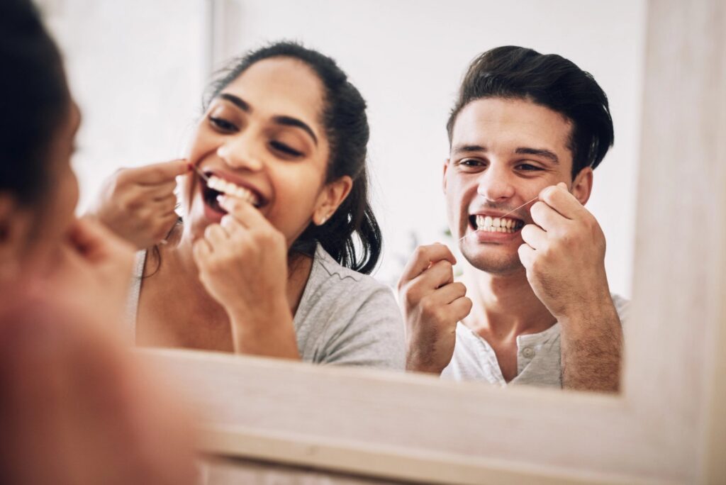 Beyond the Brush: Why Flossing Is Non-Negotiable for Your Smile a man and a woman are flossing their teeth in front of a mirror .