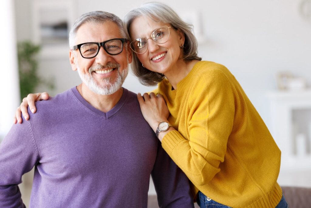Implant-Supported Dentures Vs. Traditional Dentures: Which Smile Feels More Like You? Smiling older couple with woman's arm around man.