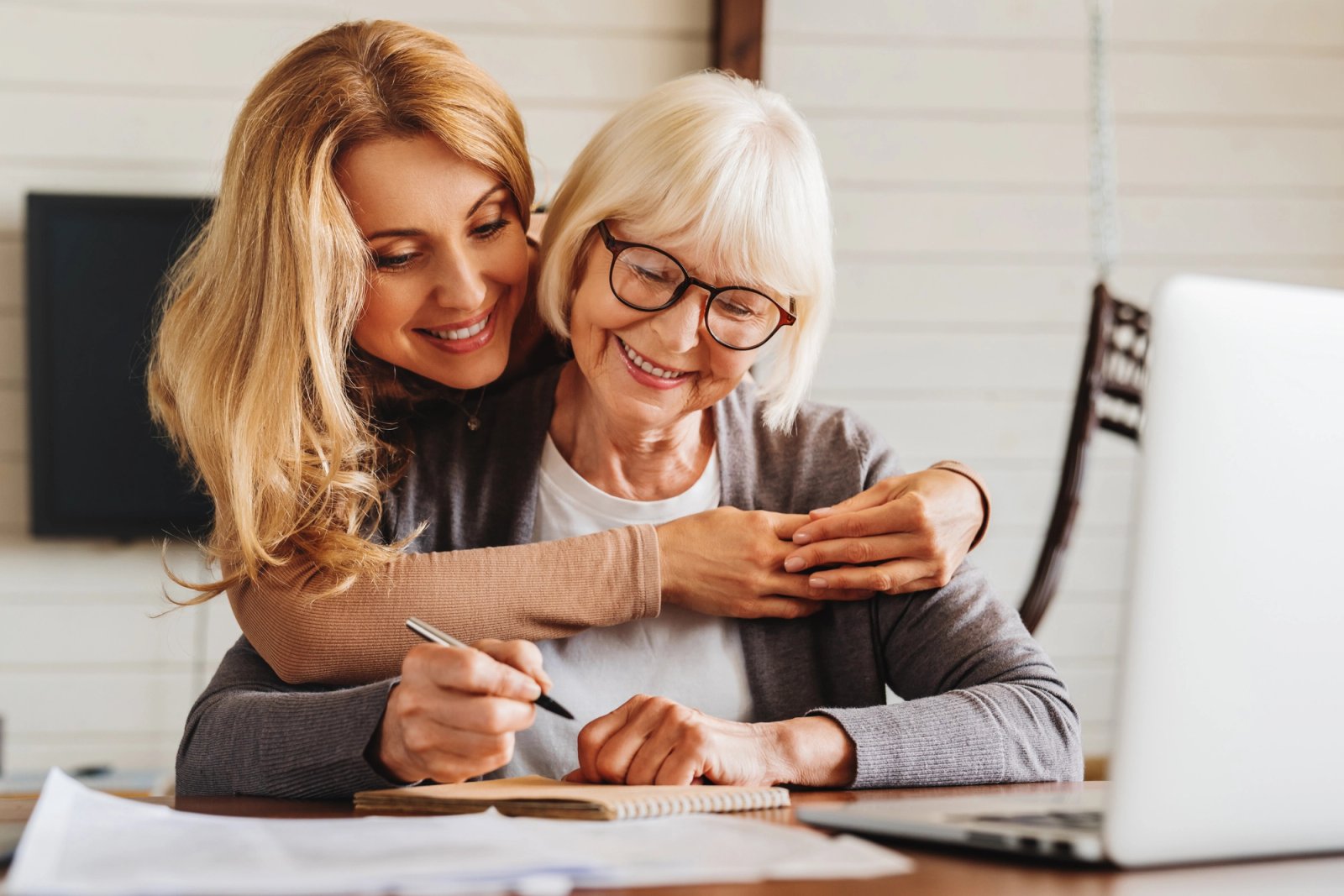 A younger woman hugs an older woman who is writing in a notebook, both smiling.